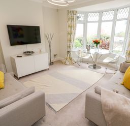 A living room with two beige sofas a round glass table with chairs a wall-mounted TV and bay windows with floral curtains at Robin Hill in Duporth near St Austell