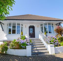 A front view of a white house with steps leading to a wooden door and flower beds on either side at Robin Hill in Duporth near St Austell
