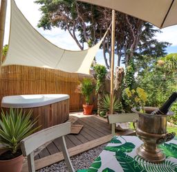 An outdoor patio area with a hot tub shaded by a white sail and a table with a bottle in an ice bucket at Roncon's Villa in Goldsithney