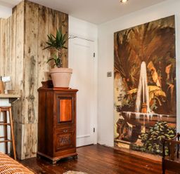 A corner of a room with wooden flooring a wooden cabinet with a potted plant a large painting of a fountain on the wall and wooden stools at Roncon's Villa in Goldsithney