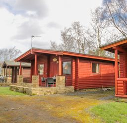 A group of red wooden cabins with stone patios and outdoor chairs in a natural setting at Ingram in Longframlington