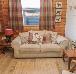 A living room with a beige sofa and cushions including one with a deer print wooden side tables with a red lamp and a plant plaid curtains and wooden walls at Ingram in Longframlington