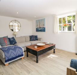 A living room with a gray sofa with blue cushions a wooden coffee table a round wall mirror a painting of sailboats and a window with flowers at The Holt in Harcombe near Sidford