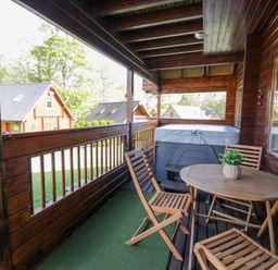 A covered wooden balcony with a round table wooden chairs a hot tub and wooden houses in the background at Sun View Lodge in Llanbedr