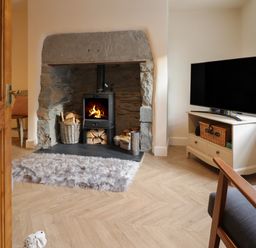 A living room with a wood burning stove in a stone fireplace with baskets of wood a TV on a stand and a chair at Bodlondeb in Tremadog