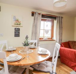 A dining area with a round wooden table set for four with white chairs next to a window and a red sofa at Lavender Cottage in Penruddock