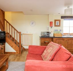 A living room with a red couch a wooden coffee table with pink flowers a television wooden stairs and a kitchen area at Lavender Cottage in Penruddock