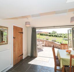 A dining area with a table covered in a green polka-dot tablecloth and wooden chairs next to open glass doors leading to a patio with outdoor furniture and garden at Lilac Cottage in Wookey Hole near Wells