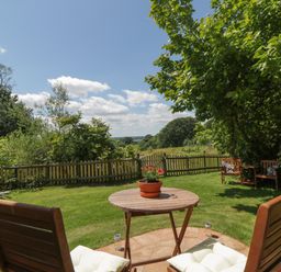 A garden with wooden chairs a round table with a flower pot on grass and a wooden fence at The Hideaway at Burrow Hill in Ottery St Mary