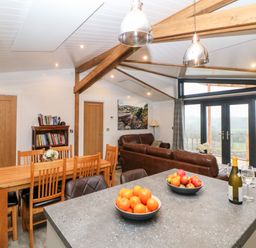 A kitchen island with bowls of fruit and a bottle of wine overlooking a dining area and living room with leather sofas and large windows at Stables Lodge in Wingerworth