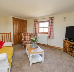 A living room with a sofa and armchair around a coffee table with flowers a television on a wooden stand and a window with plaid curtains at Bramblewood Cottage in Crookdake near Bassenthwaite