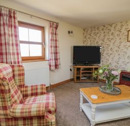 A living room with a plaid armchair a wooden coffee table with flowers a television on a stand and floral wallpaper at Bramblewood Cottage in Crookdake near Bassenthwaite