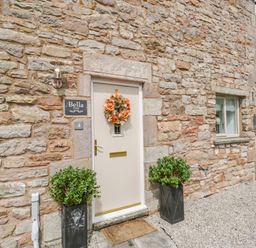 A stone exterior with a cream door decorated with a floral wreath and two potted plants at Bella Cottage Bolton near Appleby-in-Westmorland