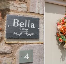 A stone wall with a plaque that reads Bella Cottage a green square plaque with number 4 and a beige door with a floral wreath at Bella Cottage in Bolton near Appleby-in-Westmorland