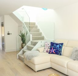 A living area with a beige sectional sofa decorated with cushions next to carpeted stairs with glass railing and a potted plant at Sail Loft in Dawlish