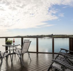A wooden deck with chairs a round table with a bottle and glasses overlooking a body of water at The Summerhouse in Y Felinheli