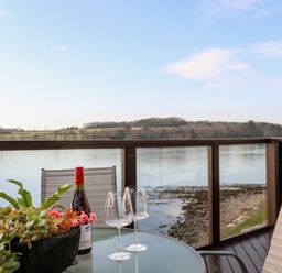 A glass table with a bottle of wine two wine glasses and a plant on a balcony overlooking water and hills at The Summerhouse in Y Felinheli