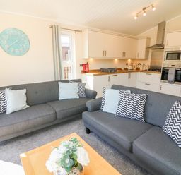 A living room with two gray sofas and cushions next to a kitchen with white cabinets at Eden Lodge - Sherwood 21 in South Lakeland Leisure Village