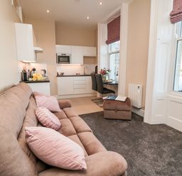 A living area with a brown sofa and pink cushions next to a small kitchen and dining table by windows at Sunset View in Whitby