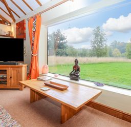 A living room with a wooden coffee table a television bookshelf and large window showing a green field at The Lodge in Hickling near Catfield
