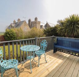 A wooden deck with a blue metal table and two chairs and a blue wooden bench with bushes and old stone buildings in the background at Sea View Lodge Eastfield Hall near Warkworth
