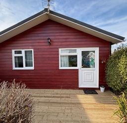 Front view of a red wooden house with two windows and a white door on a wooden deck surrounded by bushes at Trevone - Chalet 130 in St Merryn near Padstow