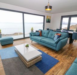 A living room with blue sofas a coffee table and large windows overlooking the sea at Machlud Haul in Llwyngwril
