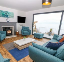 A living room with blue sofas and chairs a fireplace and a large window overlooking water at Machlud Haul in Llwyngwril