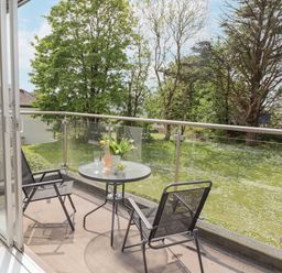 A balcony with a small round table and two chairs overlooking a grassy garden with trees at 17 Coedrath Park in Saundersfoot