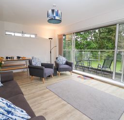 A living room with grey chairs and a sofa with patterned cushions a small dining table a floor lamp and a balcony with outdoor chairs and table at 17 Coedrath Park in Saundersfoot