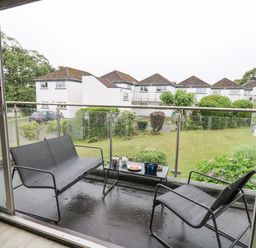 A balcony with a metal bench, chair, and table with tea set overlooking a garden and white houses at 28 Coedrath Park in Saundersfoot