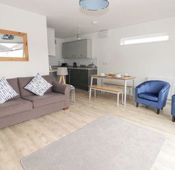 A living room with a brown sofa and two blue chairs next to a dining table with benches and a kitchen area at 26 Coedrath Park in Saundersfoot