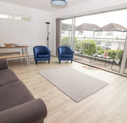 A living room with a sofa, two blue armchairs, a dining table with benches, a floor lamp, and large sliding glass doors leading to a balcony at 26 Coedrath Park in Saundersfoot