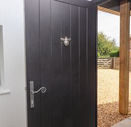 A black exterior door with a silver handle and bee decoration partially open to a gravel yard with a wooden post and fence at Bumblebee Cottage in East Nynehead near Bradford-On-Tone