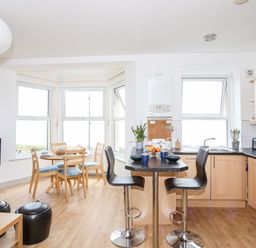 A kitchen and dining area with wooden floor a round dining table by windows and a kitchen island with bar stools at Gwel Y Mor in Pwllheli