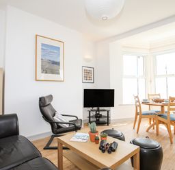A living room with a black leather sofa a black armchair a wooden coffee table with plants and binoculars and a dining table with wooden chairs at Gwel Y Mor in Pwllheli