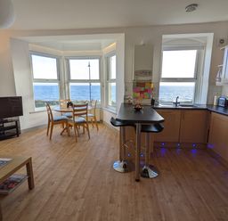 An open kitchen and dining area with a round wooden table and chairs next to large windows overlooking the ocean at Gwel Y Mor in Pwllheli