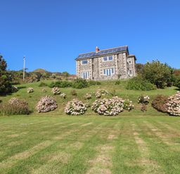 A stone house with solar panels on the roof surrounded by grass and flowering bushes at Penmarth House in Coverack