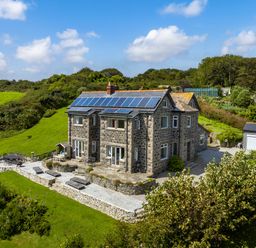 A two-story stone house with solar panels on the roof and a stone patio with outdoor furniture in a green rural area at Penmarth House in Coverack