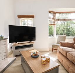 A living room with a television on a stand a coffee table with cups and decorative items a sofa and a chair near a bay window at Halewood in Bowness-On-Windermere