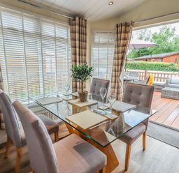 A dining area with a glass table and four chairs next to large windows and an open door leading to a patio with outdoor furniture at Black Dog Lodge Thirlmere 3 White Cross Bay near Windermere