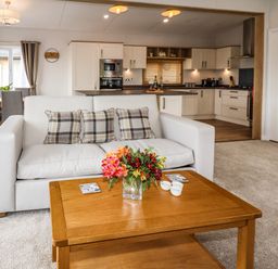 A living area with a white sofa and plaid cushions and a wooden coffee table with flowers with an open kitchen in the background at Bramblewick in Runswick Bay near Staithes