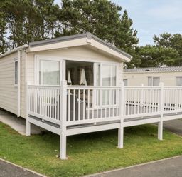 A beige mobile home with a white railing porch and sliding glass doors surrounded by trees at The Rodley Pines in Cayton