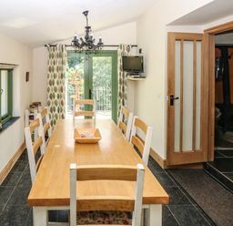 A dining room with a wooden table and six chairs near green windows and a glass door with curtains at Trewrach Cottage in Dinas Cross