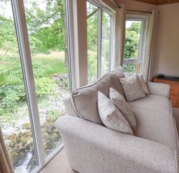 A living room with a beige sofa and cushions next to large windows showing greenery at Esthwaite Lodge in Beckside 43 Limefitt Park near Windermere