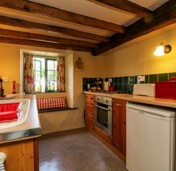 A kitchen with wooden cabinets a white fridge an oven a window with a seat below and red kitchen accessories at Bell Buoy Cottage in Morwenstow