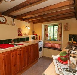 A kitchen with wooden cabinets a sink a microwave a clock and a window seat at Bell Buoy Cottage in Morwenstow