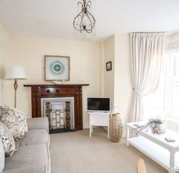 A living room with a sofa, wooden fireplace, television, floor lamp, white coffee table, and large window with curtains at Hyfrydle in Borth-Y-Gest