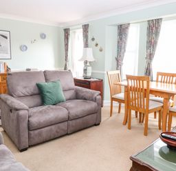 A living room with a grey sofa a wooden dining table with chairs and floral curtains at Ellan Vannin in Sheringham