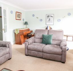 A living room with gray sofas a wooden piano a table lamp and framed pictures on the wall at Ellan Vannin in Sheringham